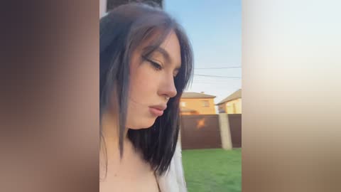 Video of a young woman with long black hair, fair skin, and a slender physique, seen from the side. She wears a white top and is standing in a suburban backyard, with a wooden fence and houses in the background.