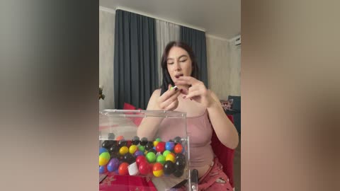 Video of a young Asian woman with light skin, dark hair, wearing a pink tank top, playing with colorful candy in a transparent container, in a cozy living room with blue curtains and light-colored walls.