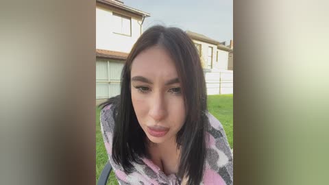 A video shows a young woman with straight black hair and light skin, wearing a pink and white checkered shirt, standing in front of a suburban house with a green lawn.