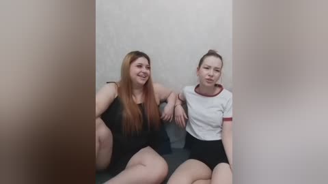 Video of two Asian women with long hair, wearing black and white sports attire, sitting close together on a black bench in a minimalistic, white-walled room.