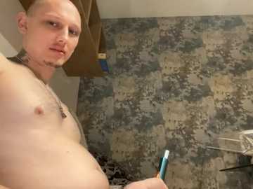 Video of a bald, fair-skinned, shirtless man with a tattooed neck holding a toothbrush in a bathroom with camouflage-patterned wallpaper.