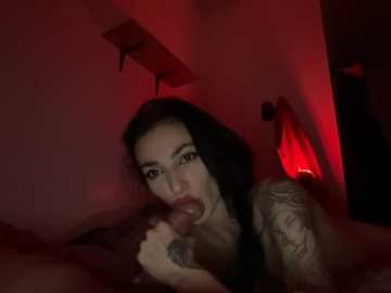 A dimly lit, grainy video of a tattooed woman with long black hair performing oral sex on a man, with a red glow illuminating the scene.