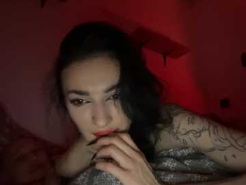 Video of a pale-skinned, tattooed woman with long, wavy black hair, wearing red lipstick, lying on a bed under red lighting. She appears pensive, resting her chin on her hand.