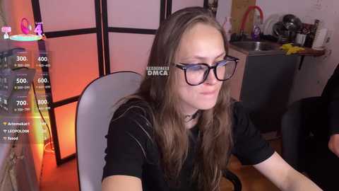 A video shows a young woman with long brown hair, wearing black-framed glasses and a black shirt, sitting at a desk in a dimly lit room with a pink and orange ambient light.