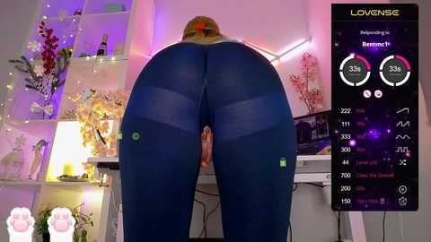 Video of a woman with fair skin, large breasts, and a curvaceous figure, wearing tight, dark blue leggings, bending over a table in a cozy, pink-lit room with floral decorations and a digital fitness tracker display.