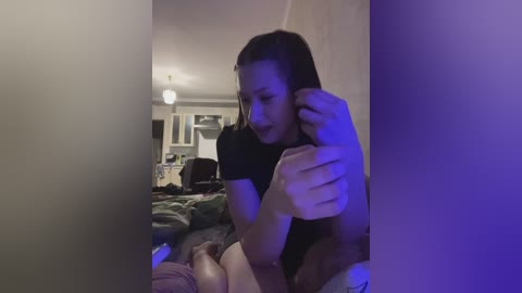 Video of a young woman with straight, dark hair, wearing a black shirt, sitting on a bed with a green blanket. Background shows a dimly lit living room with a white wall and a ceiling light.