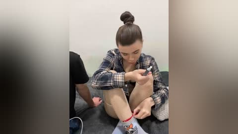 Video of a young woman with dark hair in a bun, sitting on a grey couch, tying her shoe, wearing a plaid shirt, in a minimalist room with white walls.