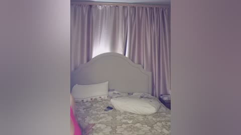 Video of a cozy bedroom with a white, curved headboard, beige floral bedspread, and matching pillows. Soft pink curtains cover the window, creating a serene, feminine ambiance.