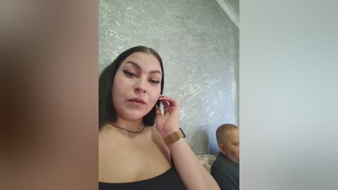 Video of a young, light-skinned woman with straight black hair, wearing a black off-shoulder top, and a gold watch, sitting on a couch. In the background, a baby sleeps.