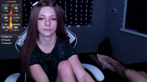 A video depicts a young woman with fair skin, long brown hair, and pink eyeshadow, seated on a gaming chair in a dimly lit room. Her expression is neutral. A TV screen shows a \"Loweing\" countdown.