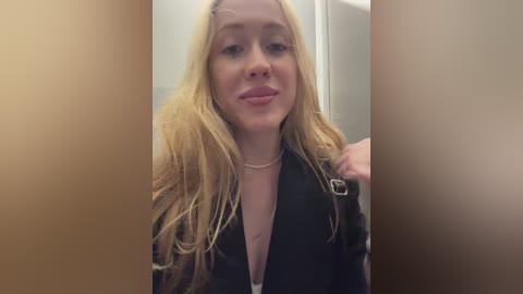 A video of a young woman with long, wavy blonde hair and fair skin, wearing a black jacket and a silver necklace. She has a slight smile. Background is blurred.