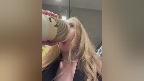 Video of a blonde woman in a black jacket, drinking from a beige coffee cup with a beige lid, in a modern, dimly lit room with a potted plant.