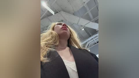 Video of a blonde woman with long hair in a black blazer, wearing a white shirt, standing in a modern, industrial building with white ceilings and exposed pipes.
