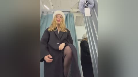 Video of a blonde woman in a navy coat and white hat, standing in a dressing room with a full-length mirror.