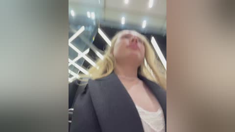 A blurry video of a blonde woman in a grey blazer, leaning back in a modern, bright room with a glass and metal interior.