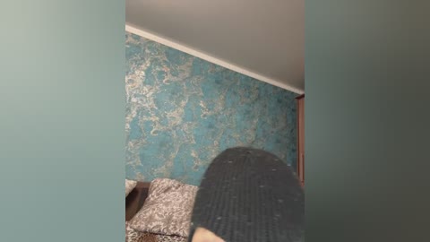 Video of a teal, textured wallpapered wall with a white ceiling. A person's legs are visible in the foreground, wearing brown and gray patterned socks.