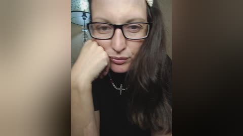 Video of a Caucasian woman with long brown hair, glasses, and a black T-shirt, leaning on her hand, looking serious. Background features a beige wall and a lampshade.