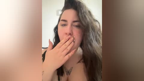 A video of a young woman with long, wavy brown hair, partially covering her face, with a butterfly tattoo on her chest and a small tattoo on her hand. She's biting her finger, in a casual, indoor setting.