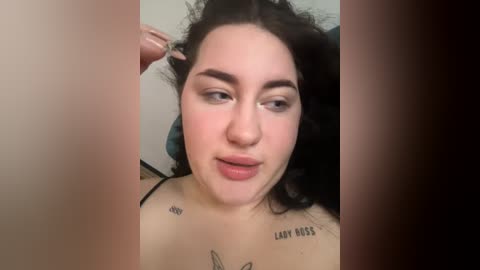 Video of a young woman with curly black hair, fair skin, and prominent eyebrows, wearing a black top, touching her hair. She has tattoos on her chest. Background is blurred.