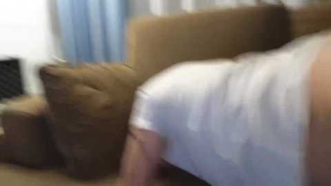 Video of a person with short, dark hair lying on a beige sofa, wearing a white T-shirt, in a dimly lit room with blue curtains in the background.