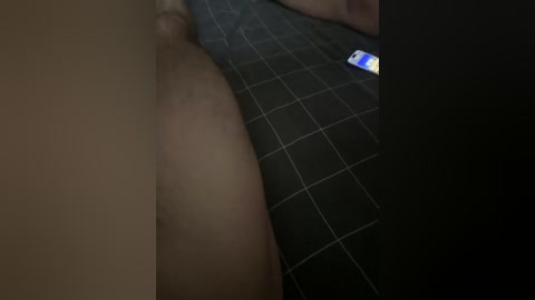 A dimly lit video showing a close-up of a person's lower torso and legs on a black tiled floor. The skin tone is light, and a smartphone with a blue light is visible in the background.