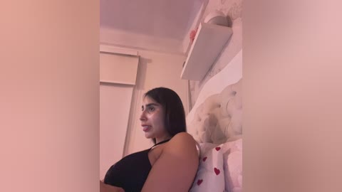 Video of a young woman with long black hair, wearing a black tank top, sitting on a bed with a heart-patterned pillow. She looks serious. Background shows a white wall and a window with a closed blind.