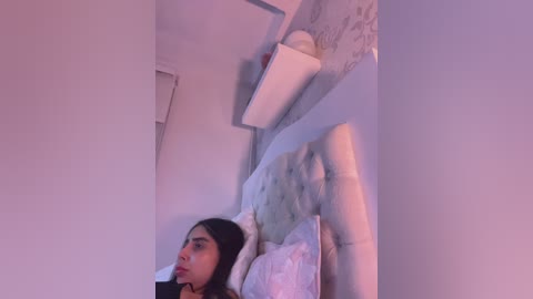 Video of a young woman with medium brown skin and long black hair lying on a white tufted bed in a dimly lit room with purple lighting, surrounded by white walls and a wall-mounted shelf.