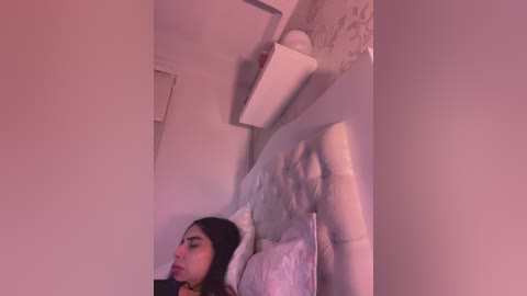 Video of a young woman with dark hair, lying on a bed, partially obscured by a white sheet. The room is dimly lit, with a white shelf and a light fixture visible in the background.