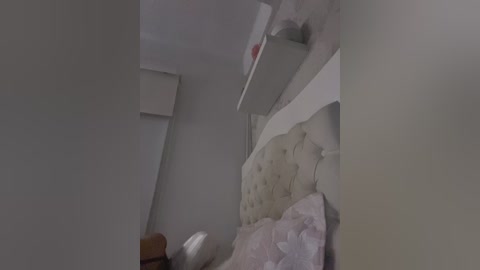 Video of a small, dimly lit bedroom with a cream-colored, tufted headboard, a white shelf, and a beige wall. The bed has a white sheet and a pink, frilly pillow. The room appears cluttered with objects.