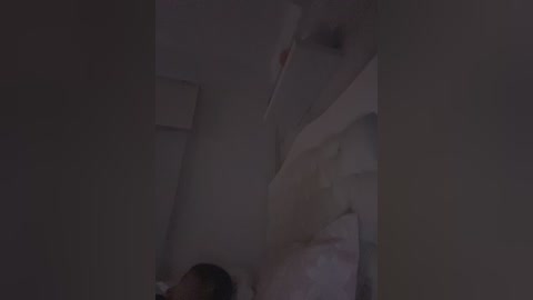 A dimly lit video captures a man in a white shirt, possibly sleeping, in a cluttered, messy room with a bed and partially visible wall. The scene is dimly lit, creating a moody, somewhat eerie atmosphere.