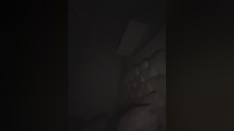 A dimly lit, close-up video of a person's face, partially obscured by shadows. The subject appears to be a male with short, dark hair and a beard. The background is indistinct and dark, emphasizing the subject's face.