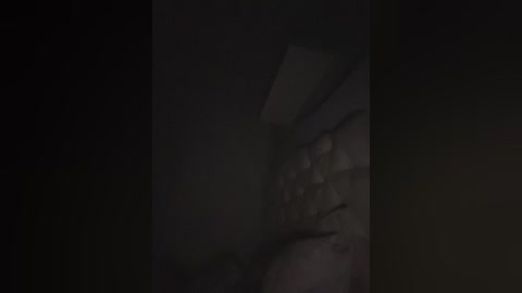 A dimly lit video showing a close-up of a person's face, partially obscured by shadows, with visible facial features and a subtle, dark background.