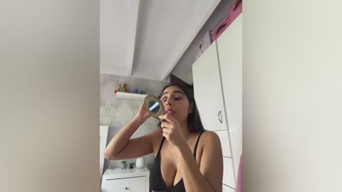 A video of a young woman with medium skin tone and long black hair, wearing a black spaghetti-strap dress, applying lipstick in a bathroom with white cabinets and a pink towel.