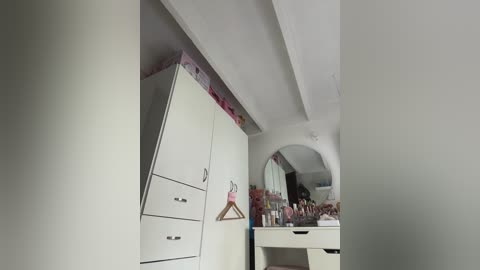 Video of a minimalist, white-walled bedroom with a white wardrobe, a vanity table with makeup, and a hanging mirror.