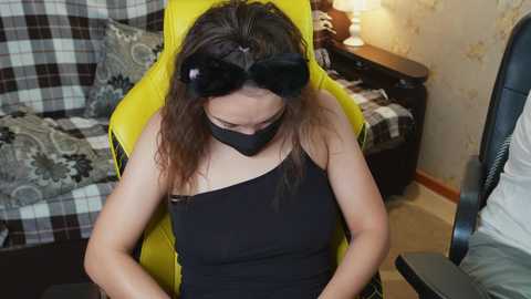Video of a young woman with long brown hair, wearing a black blindfold, black mask, and sleeveless black top, seated in a bright yellow gaming chair in a modestly furnished room with patterned curtains and a wooden side table.
