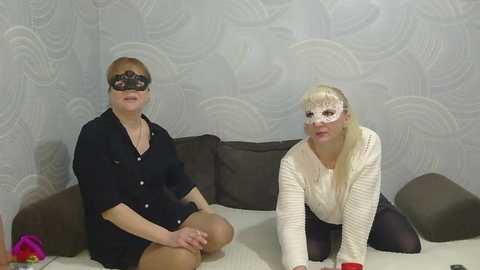 Video of two women dressed in black and white costumes, sitting on a sofa with a floral-patterned wall. The woman in black wears a lace mask, while the woman in white has a lacy mask and a white knit cardigan.