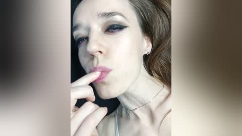 A video of a young, fair-skinned woman with auburn hair, wearing dark eyeliner, kissing her fingers while gazing seductively. She has a delicate silver necklace and is wearing a light-colored top. The background is blurred and neutral-toned.