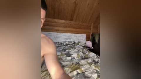Video of a shirtless man with light skin and short dark hair, lying on a bed with a patterned duvet, in a wooden attic room with a slanted ceiling.