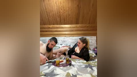 Video of a man and woman, both topless, lying on a bed with a floral-patterned sheet, sharing a drink in a rustic wooden cabin.