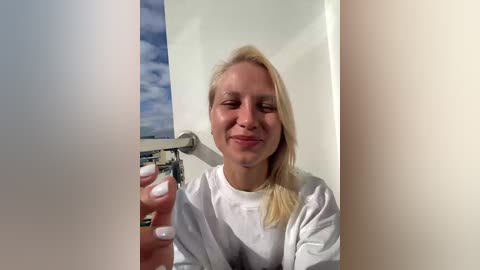 Video of a blonde woman with fair skin, wearing a white shirt, smiling and taking a selfie through a rectangular frame, with a clear blue sky and cloud background visible.