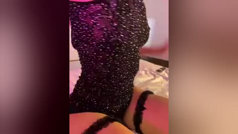 Video of a woman in a black, sparkly, sequined dress, sitting on a bed with pink sheets. Her legs are crossed, and her breasts are partially visible under the dress. The background is blurred and dimly lit.