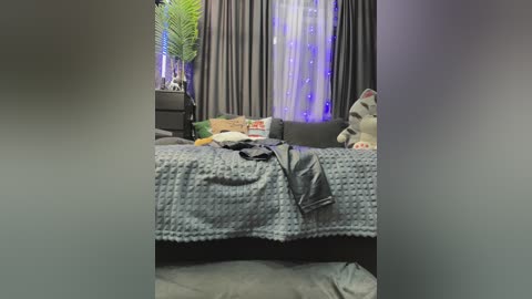 A blurry video of a dimly lit, messy bedroom with a gray quilt, a stuffed toy, and a black couch under dark curtains.