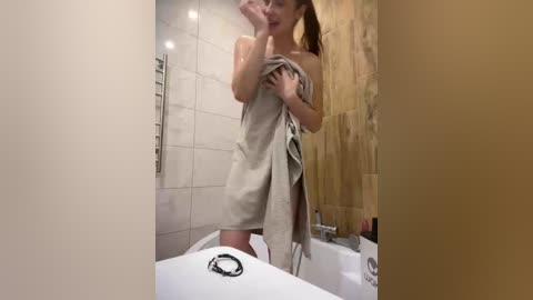 Video of a woman in a bathtub, holding a towel, wearing a beige robe, in a beige-tiled bathroom with a shower curtain.