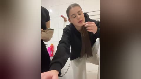 Video of a young woman with long brown hair, wearing a black jacket and white pants, leaning against a white tiled wall, with blurred figures in the background.