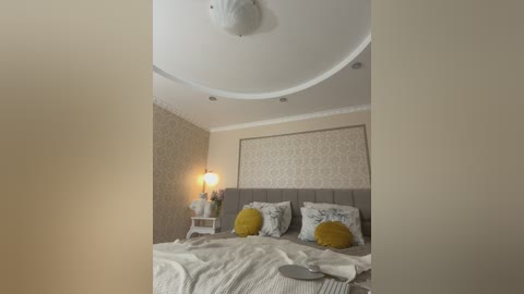 Video of a cozy bedroom with beige walls, a tufted gray headboard, two yellow pillows, a white nightstand, and a round ceiling light.
