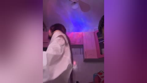 A blurred video of a woman in a white robe standing in a dimly lit kitchen with purple and blue lighting, pink towels hanging, and a ceiling fan.