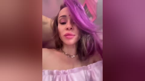 A video of a young woman with light skin, wearing a white off-shoulder top, and vibrant purple hair, posing with a relaxed expression against a blurred pink background.