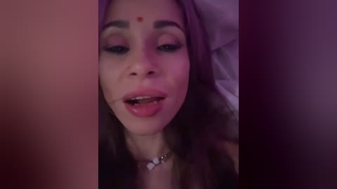 Video of a young woman with light brown skin, long brown hair, and a red bindi on her forehead. She is lying down, eyes half-open, wearing a necklace with white beads. The background is dimly lit, suggesting an intimate, possibly nighttime setting.