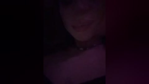 A dimly lit, blurry video of a person with short, dark hair, wearing a black choker necklace. The background is dark and indistinct, with a reddish hue on the right side.