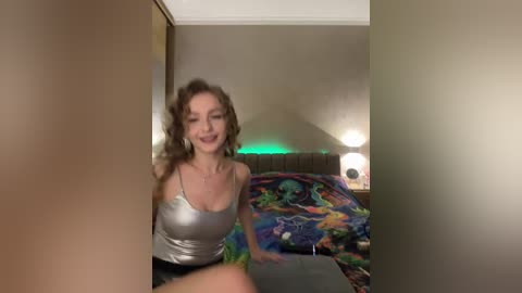 A video of a smiling, light-skinned woman with curly brown hair, wearing a silver tank top and black shorts, sitting on a colorful, patterned bed in a dimly lit room.